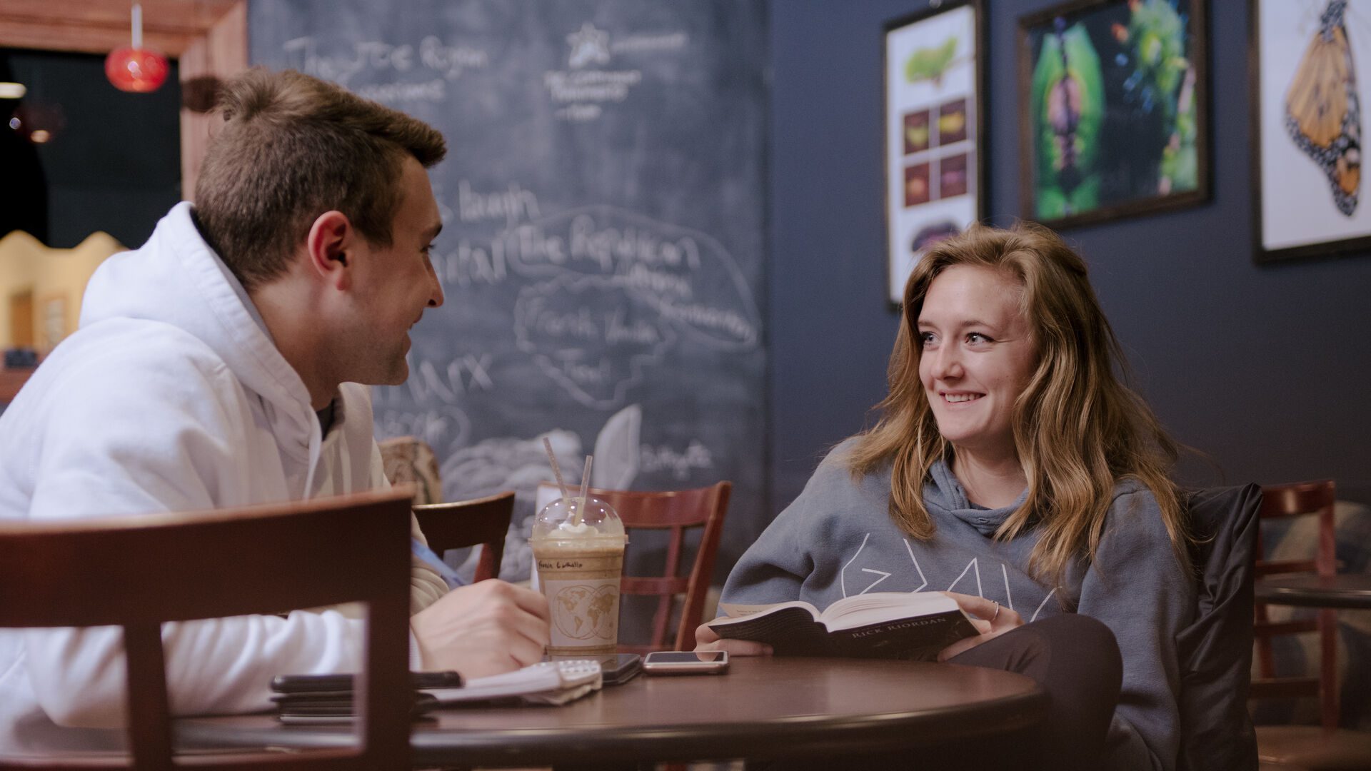 Two ����ר�� students sitting at table in Java 101 reading and talking.