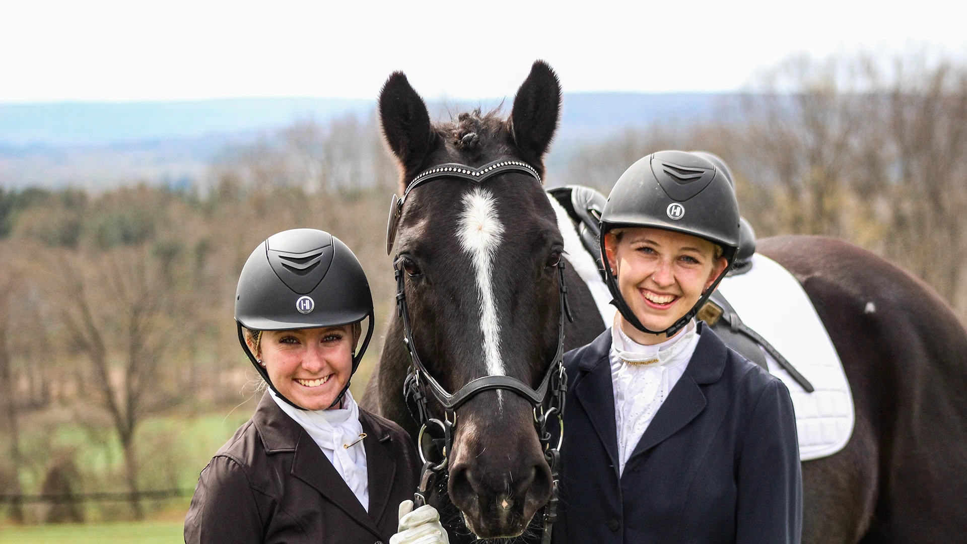 Two ����ר�� equestrian students with helmets and show clothes standing next to black lesson horse outside.