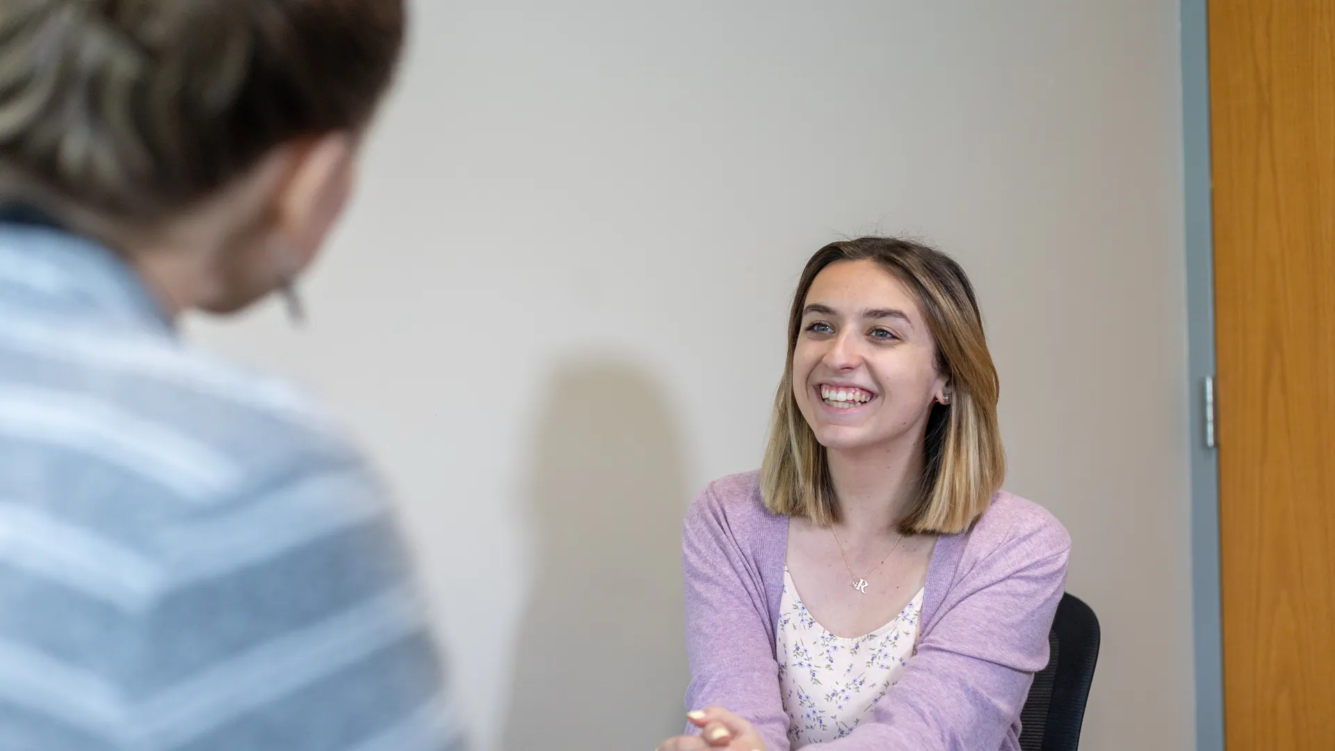 Houghton student sitting in interview room smiling and wearing a lavender sweater.