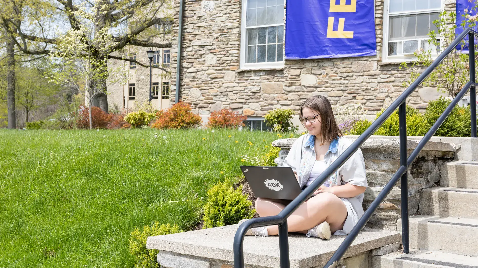 Houghton Universitystudent sitting by stairs working on laptop outside.