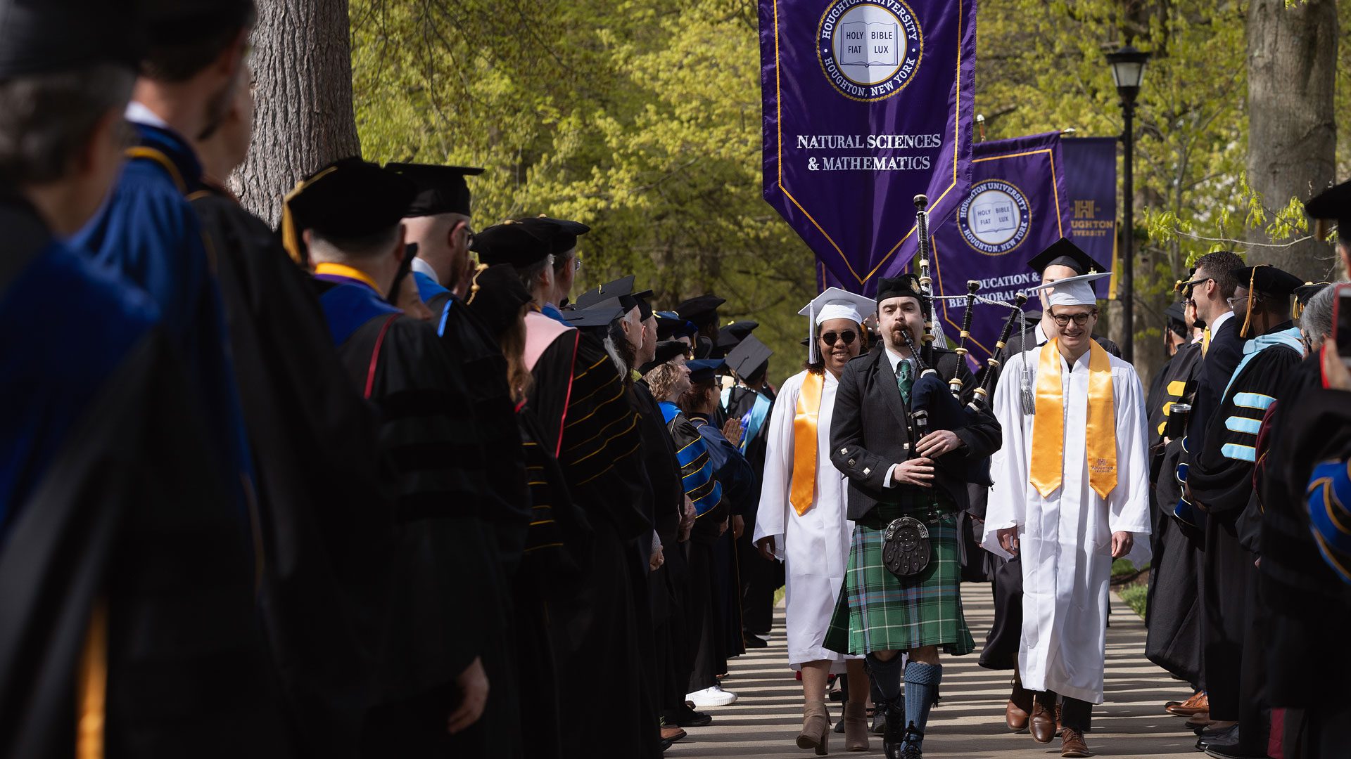 Houghton Universitystudents walking down sidewalk with bagpipers during commencement.