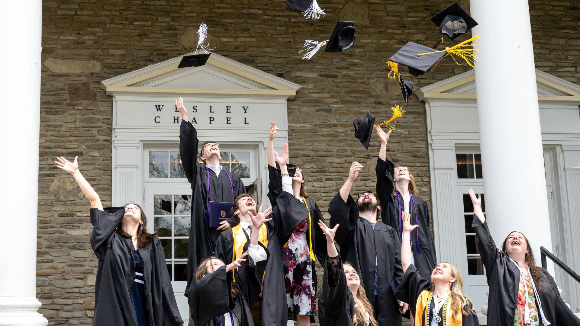 Houghton graduates standing on the Wesley Chapel steps throwing up their grad caps.
