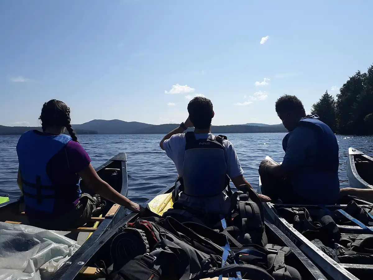 Three ����ר�� Highlander Wilderness Adventure students on canoes looking over lake.