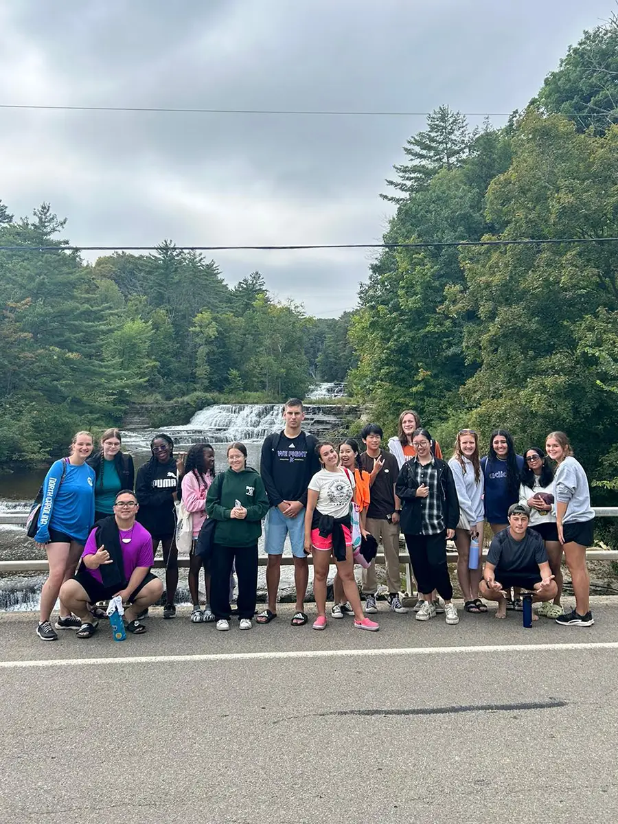 Houghton Universitygroup of interconnect students standing on road in front of waterfall.