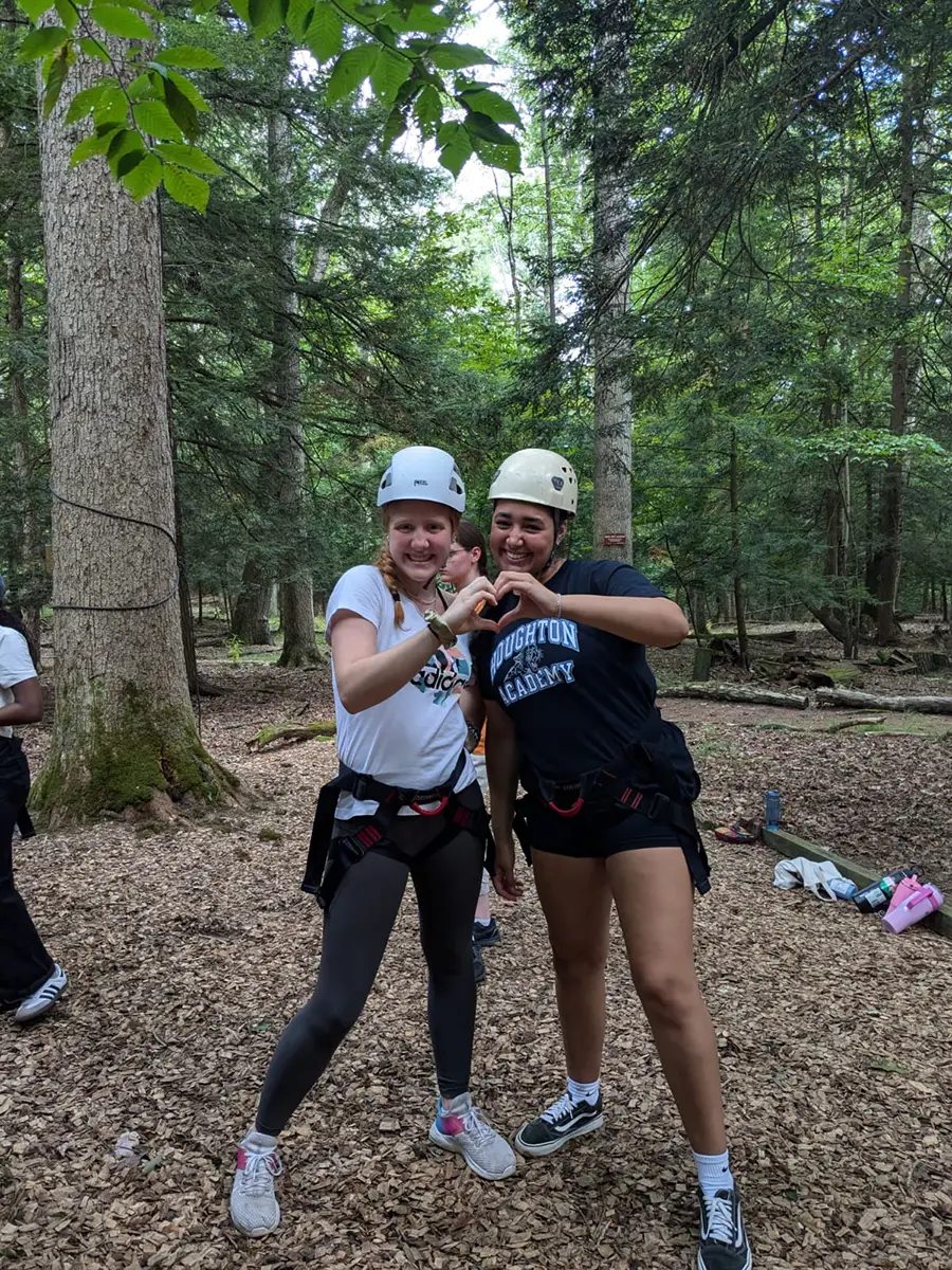 Two ����ר�� interconnect students wearing helmets on ropes course smiling and making heart with hands.
