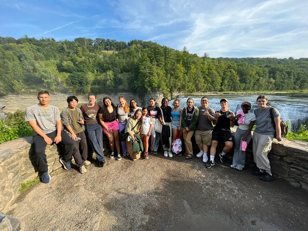 Houghton Universityinterconnect students standing on by stonewall in front of waterfall.