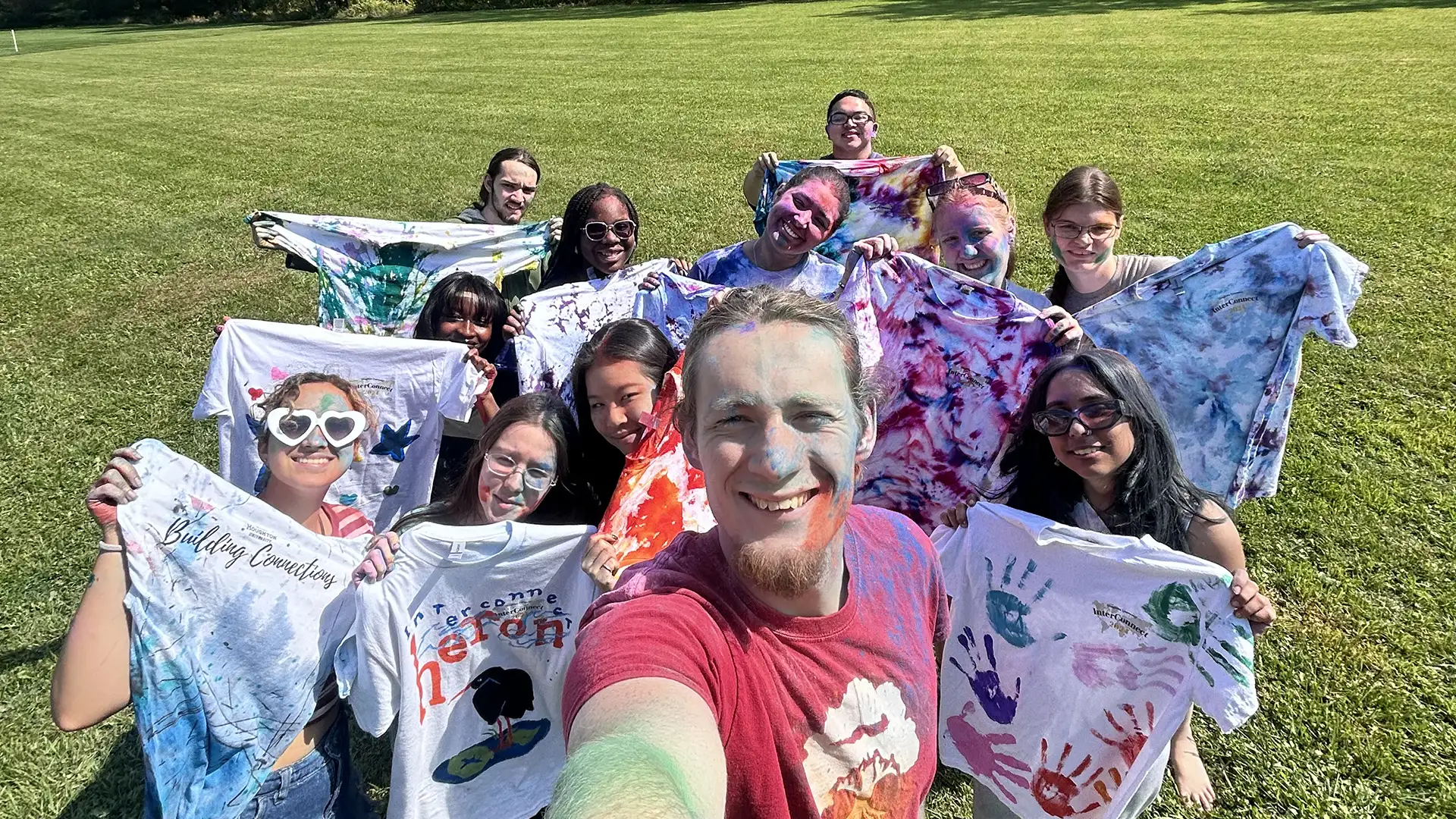 Houghton Universityinterconnect students standing together covered in powdered color holding tie dye shirts.