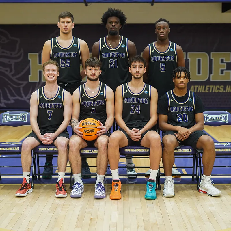 Houghton UniversityMens Basketball team sitting on the court smiling.