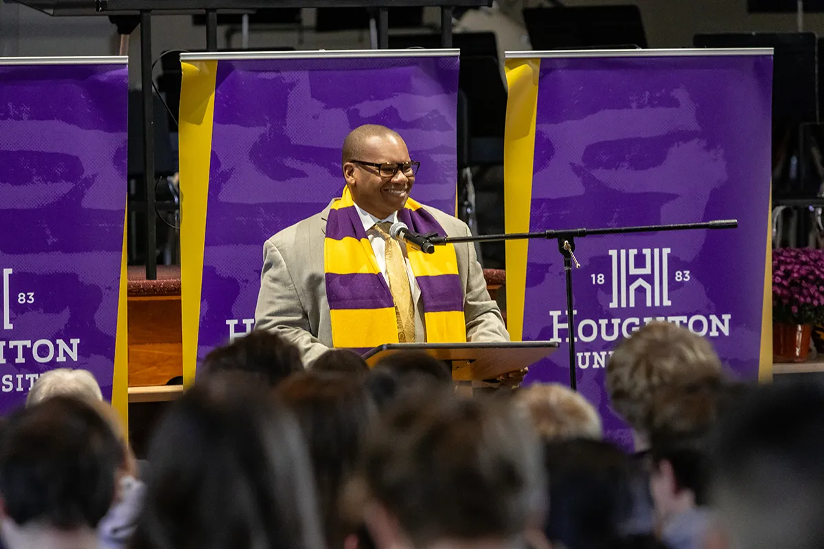 Houghton Universitypresident Wayne Lewis standing at podium wearing purple and gold scarf.