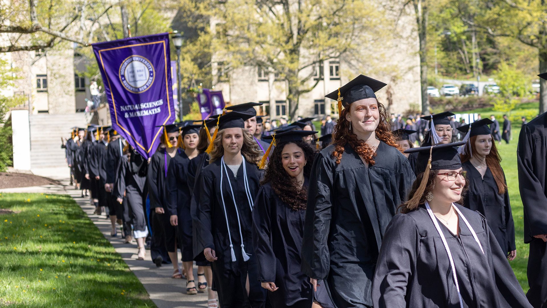 Graduates in black caps and gowns processing in a line across a grassy area. A purple banner reading 'Natural Sciences & Mathematics' is carried by students in the procession. The scene is set on a sunny day with buildings and trees in the background.
