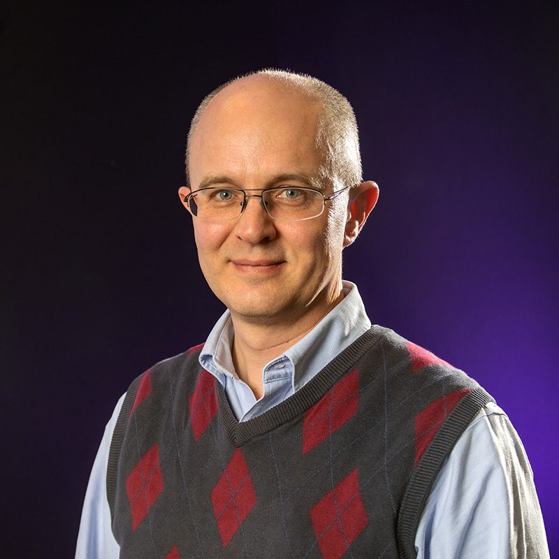 Peter Meilaender with blue button up shirt and plaid vest standing in front of purple backdrop.