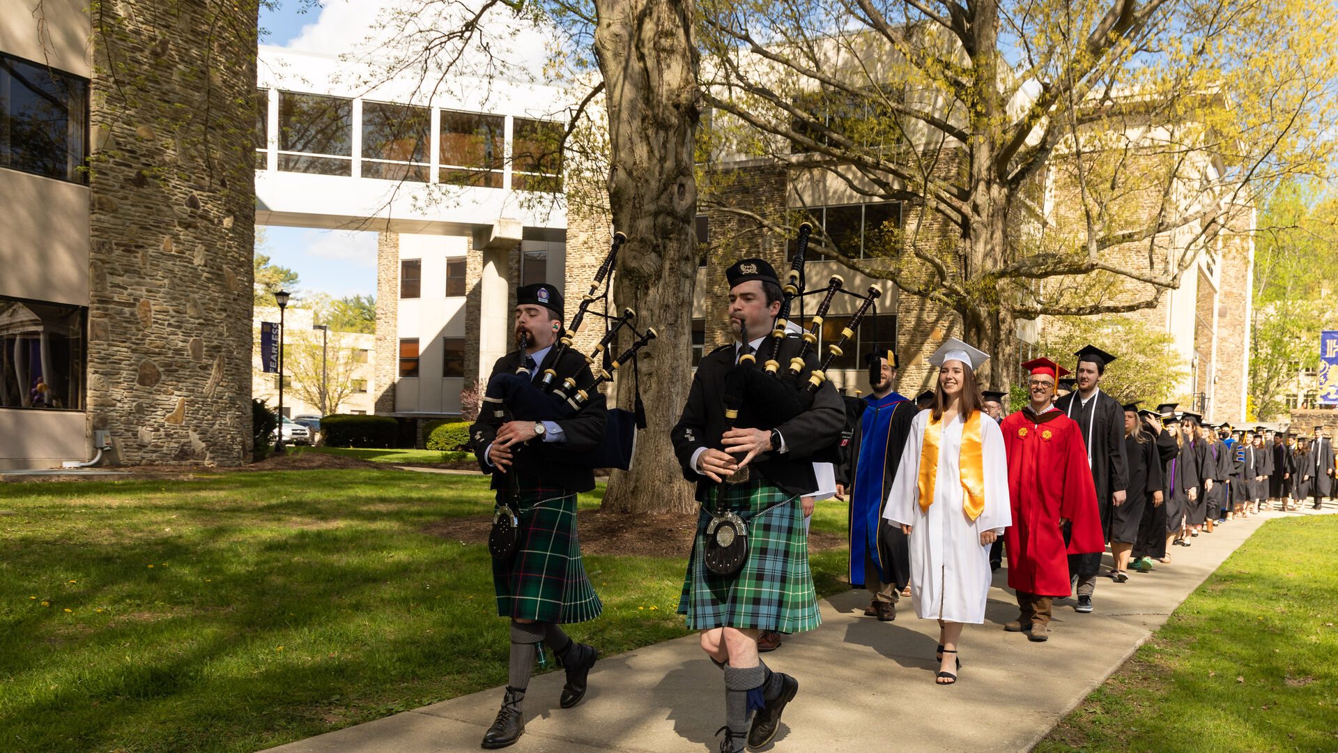 Bagpipers and ���Ͽ� graduates walking down campus path during graduation ceremony.