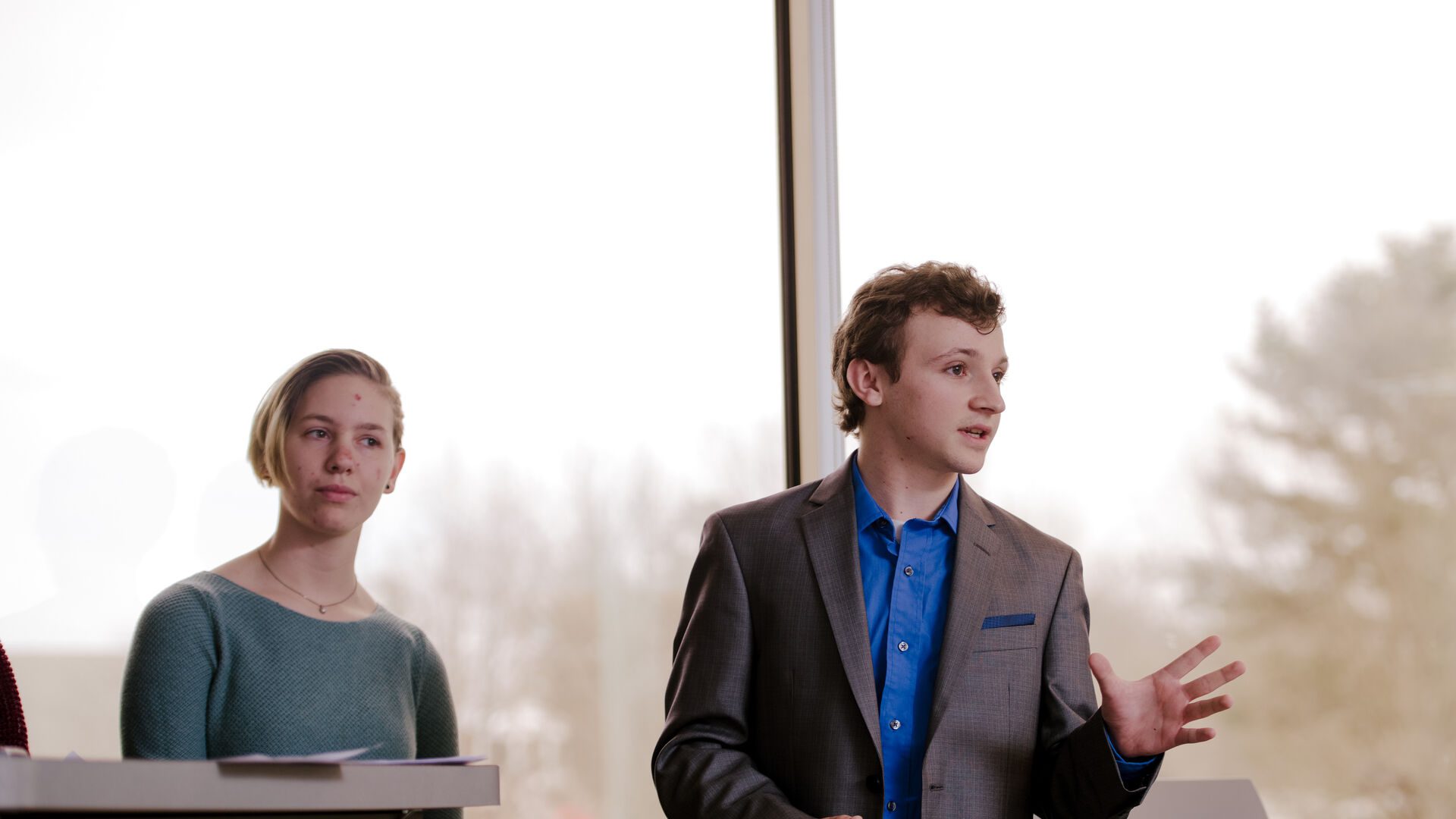 Student presenting in front of the class, wearing a blue shirt and suit coat.