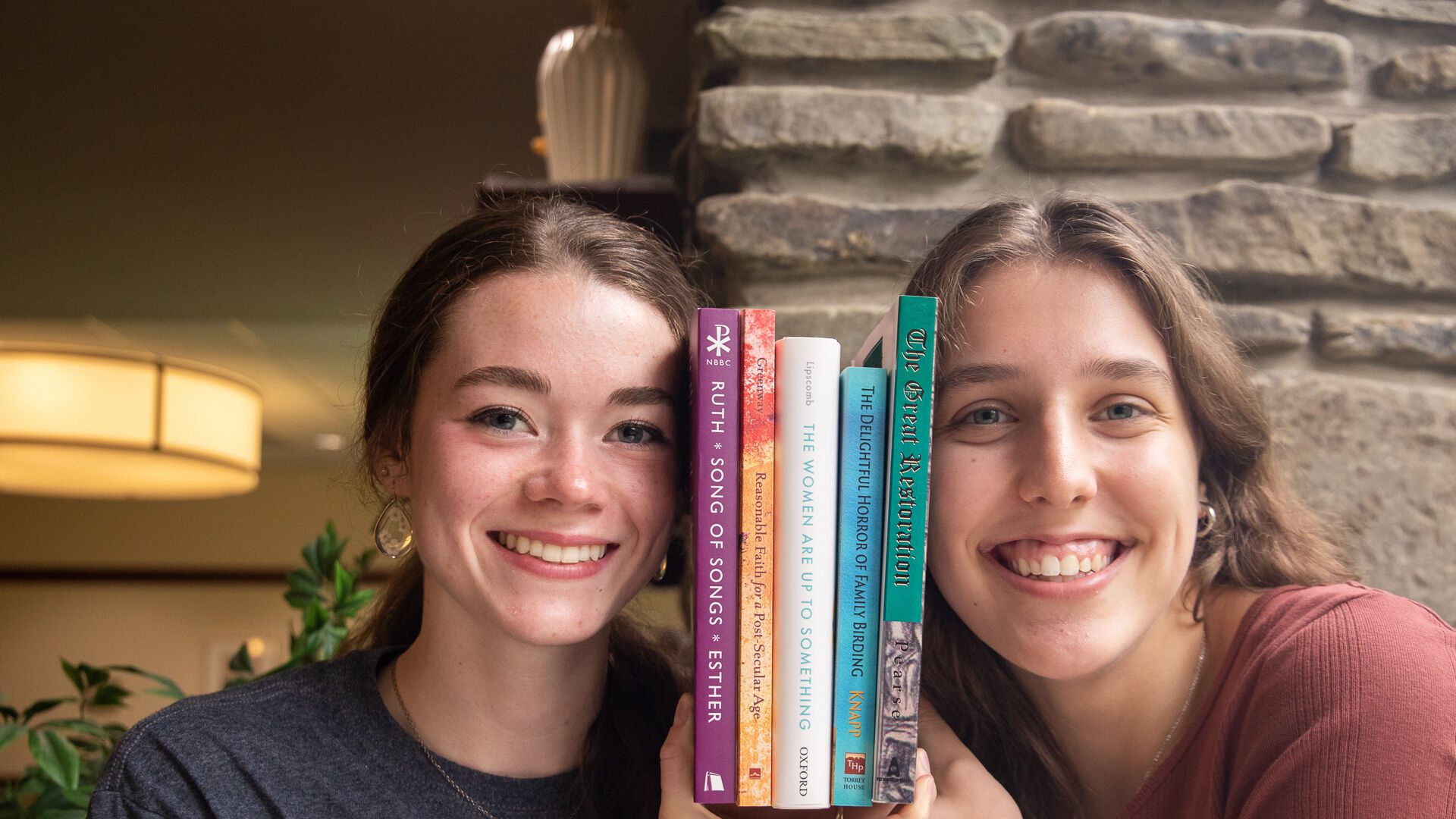 Two students smiling with a small stack of books between their faces.