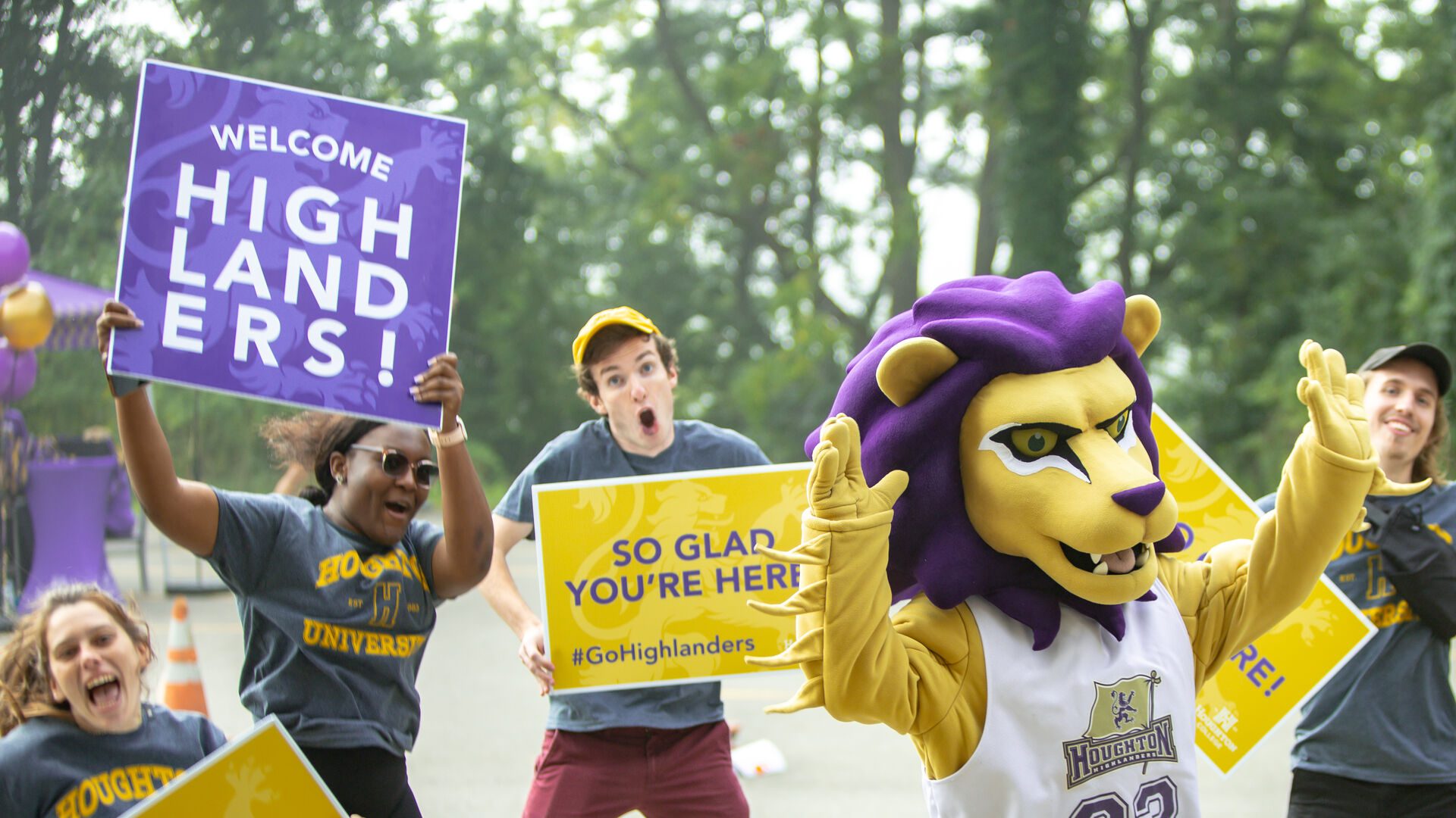 Luckey the lion mascot standing with his arms raised with a group of students jumping in the air.