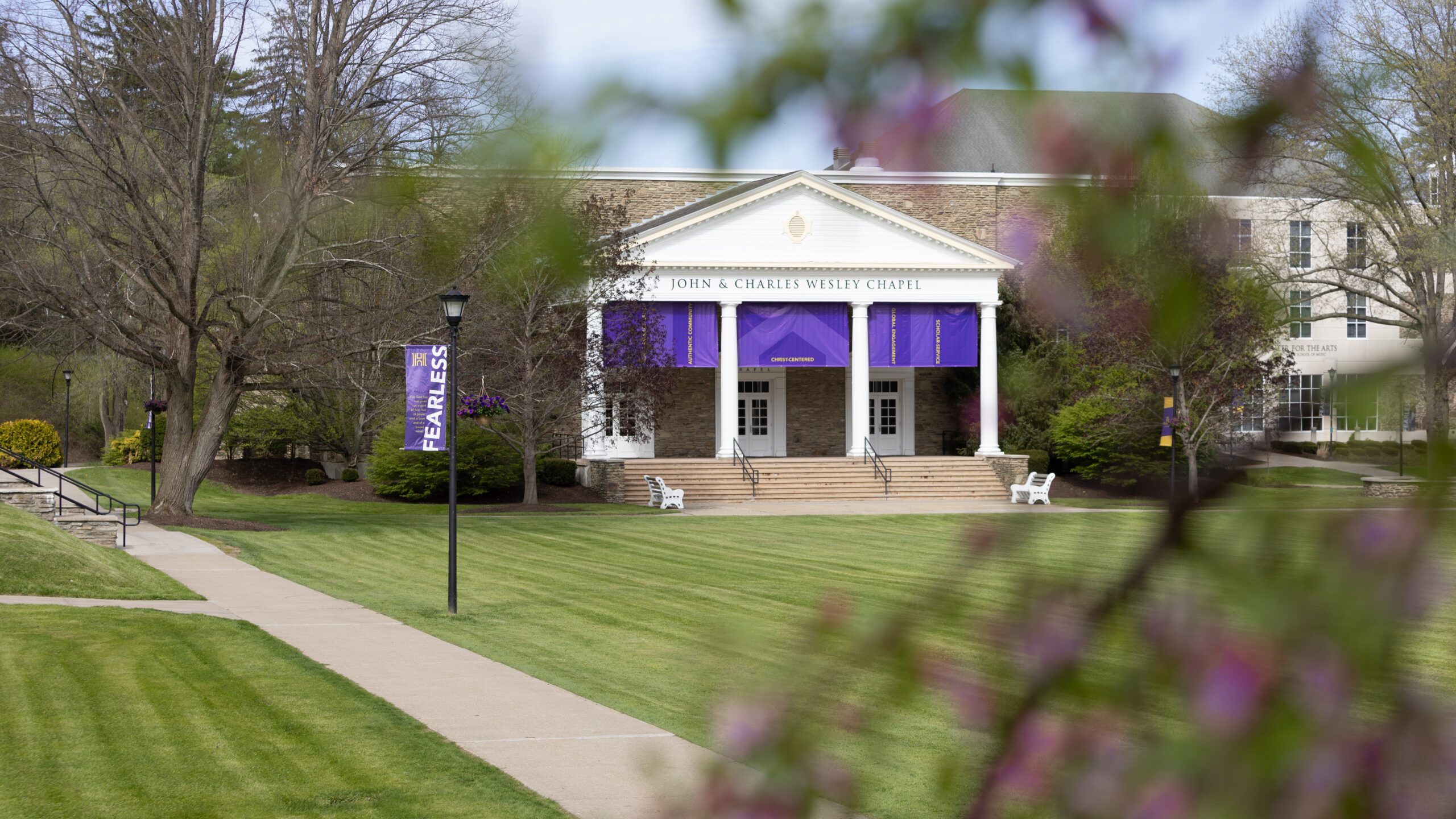 Photo of John and Charles Wesley Chapel in the springtime with flowering branches in the foreground, green grass and blue skies. Banner that says 