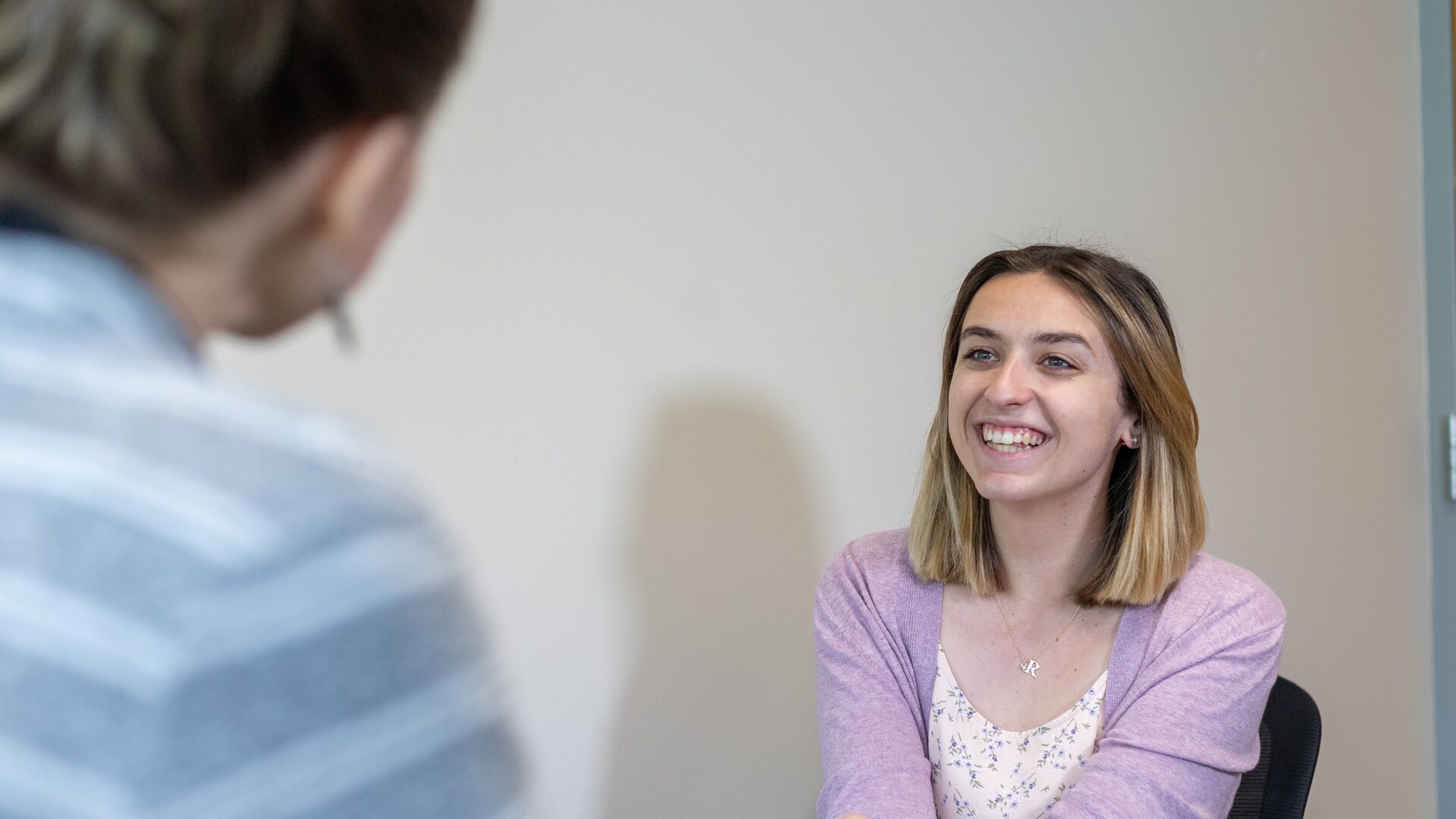 Houghton student sitting in interview room smiling and wearing a lavender sweater.