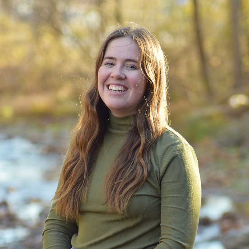 Mary Vandenbosch standing outside near creek wearing sage turtle neck.