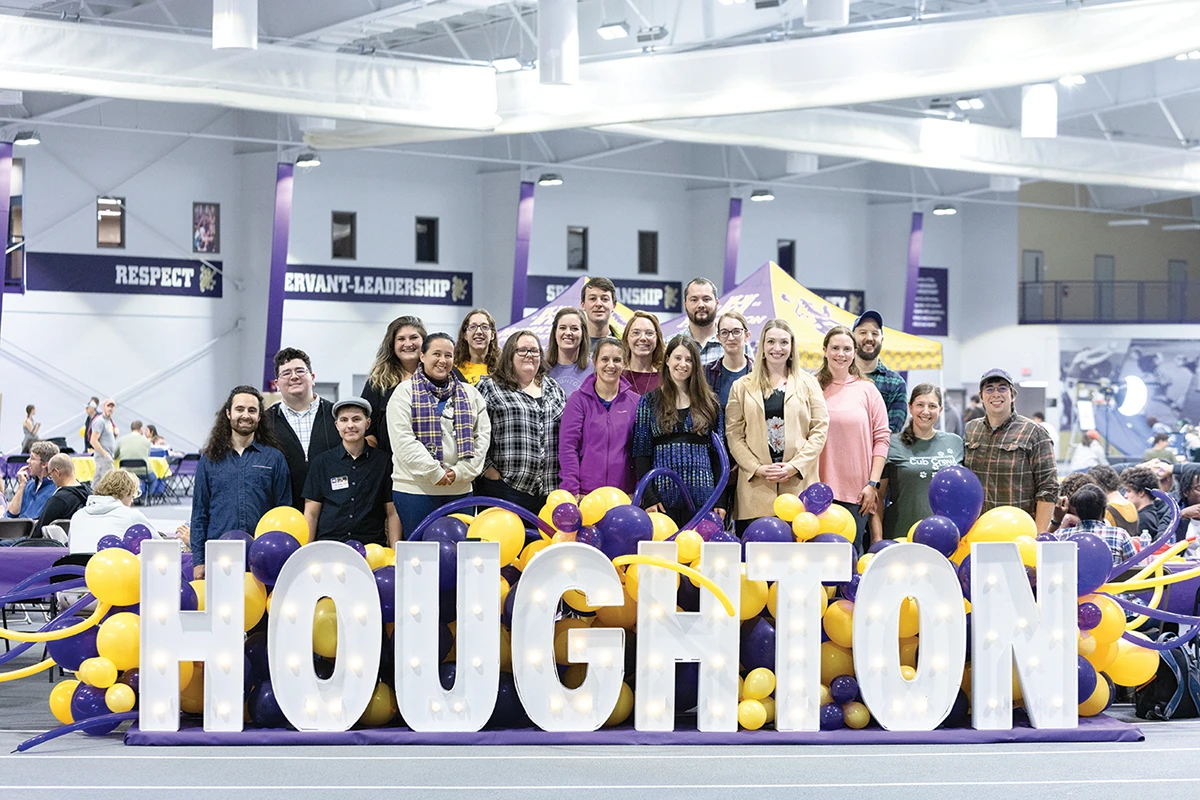 Group of Houghton alumni standing together with large Houghton light up sign in gym.