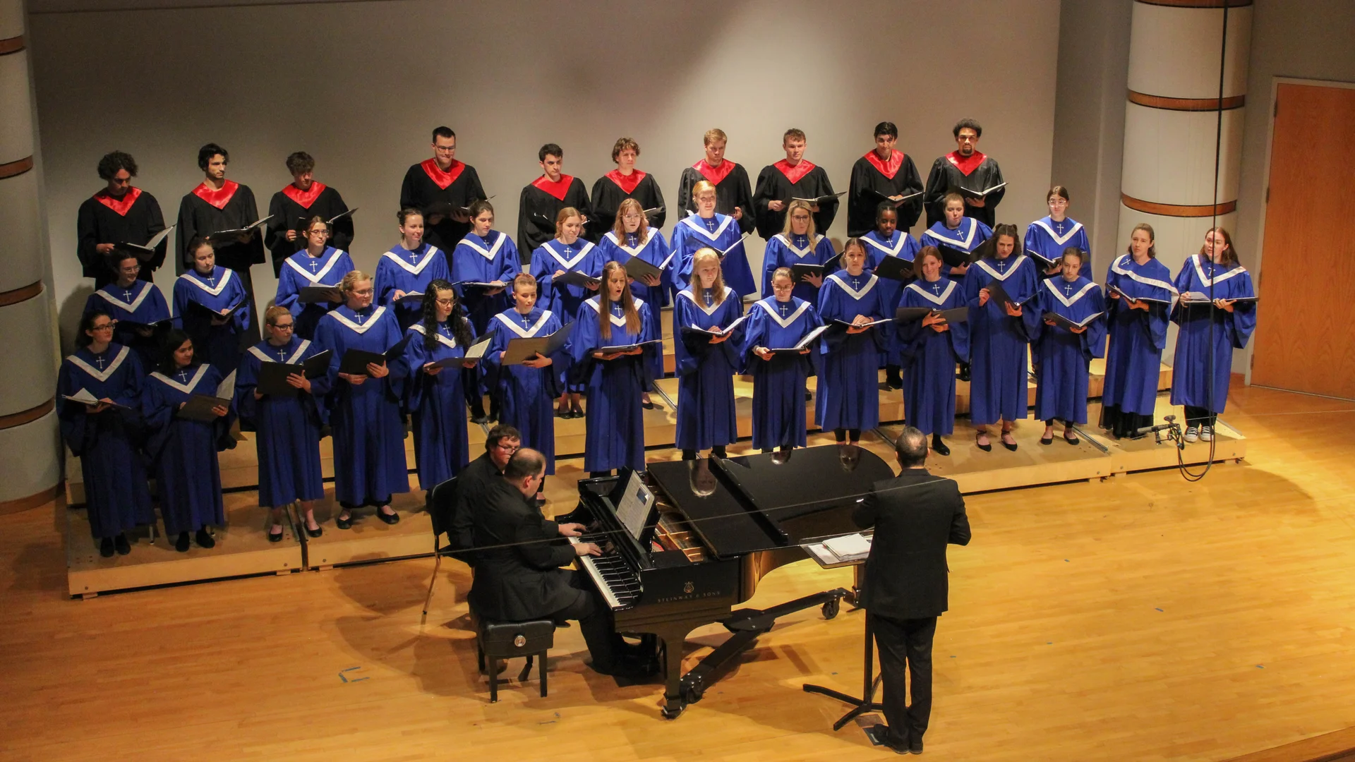 Houghton choir singing on stage with piano and conductor in traditional robes.