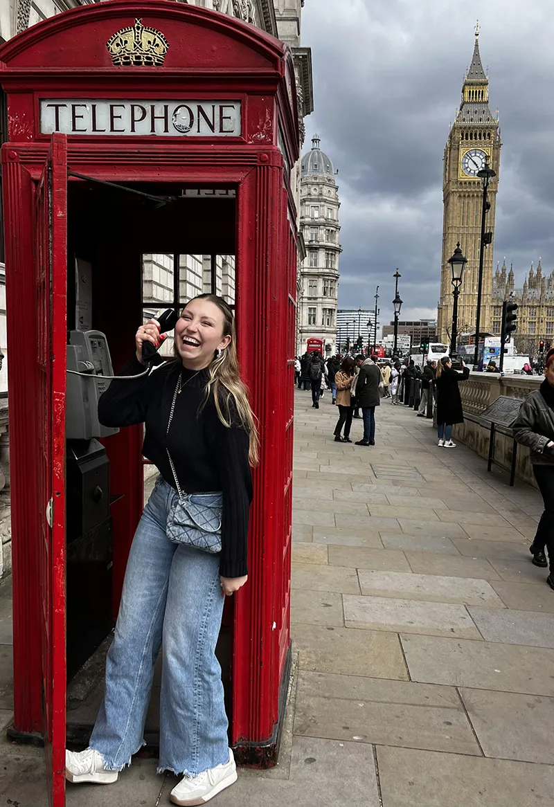 Houghton honor's student standing in red telephone booth in London with Big Ben in the background.