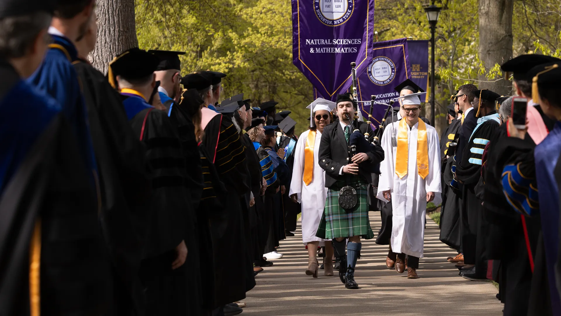 Bagpiper followed by Houghton graduates walking down path on campus quad.
