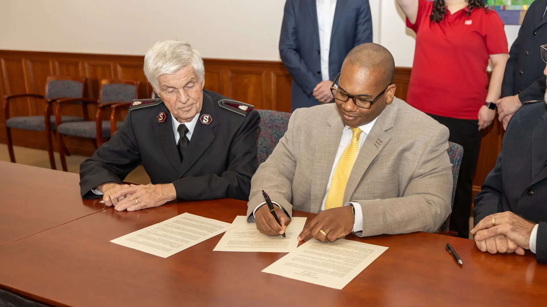 ���Ͽ� President Wayne Lewis signing document on table next to Salvation Army administrator.